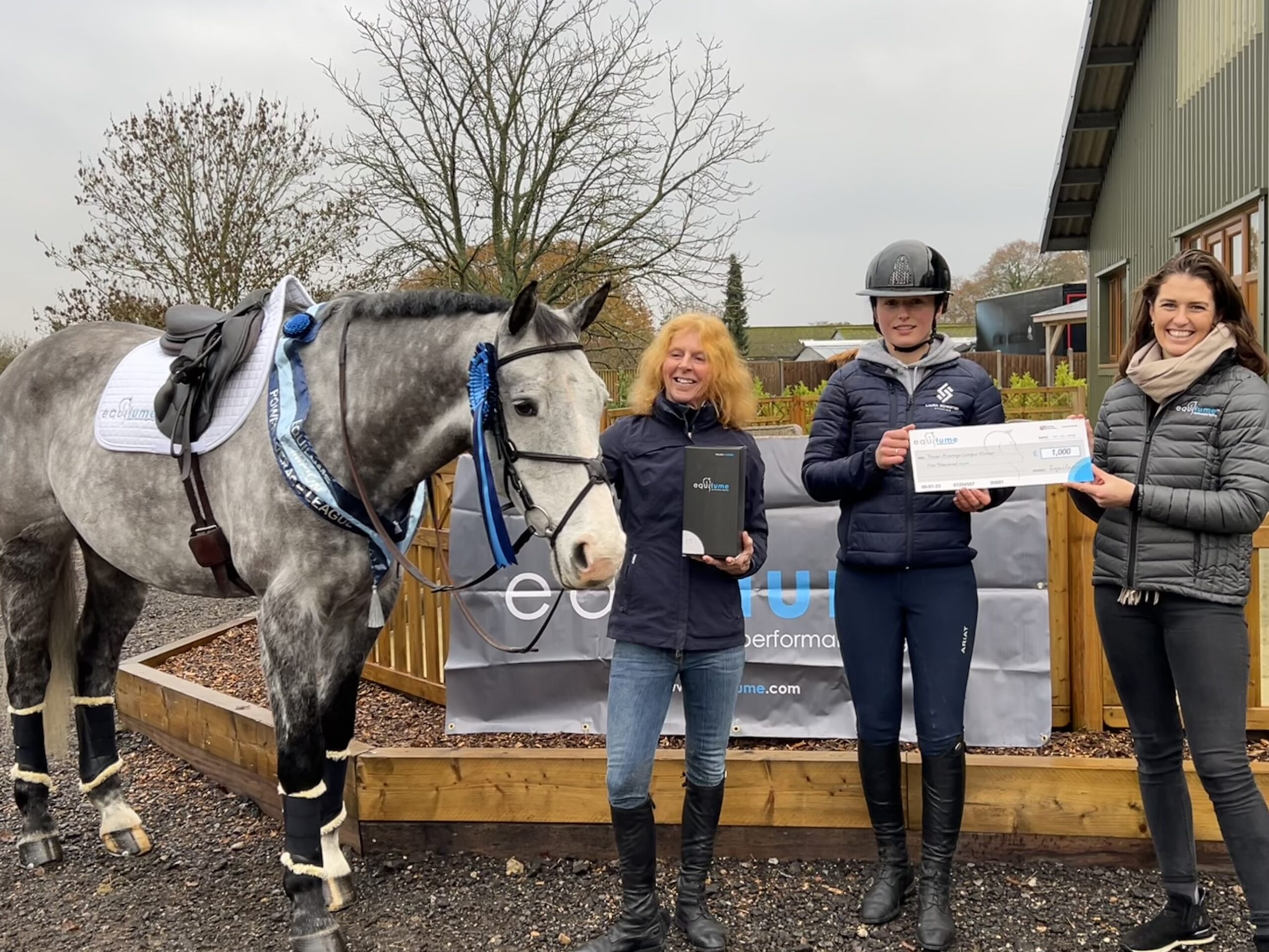 Grey horse wearing a saddle and bridle and his prize of a sash and a rosette on his head. Standing beside his owner Katie, rider Laura and Equilume marketing manager Alyssa O'Neill