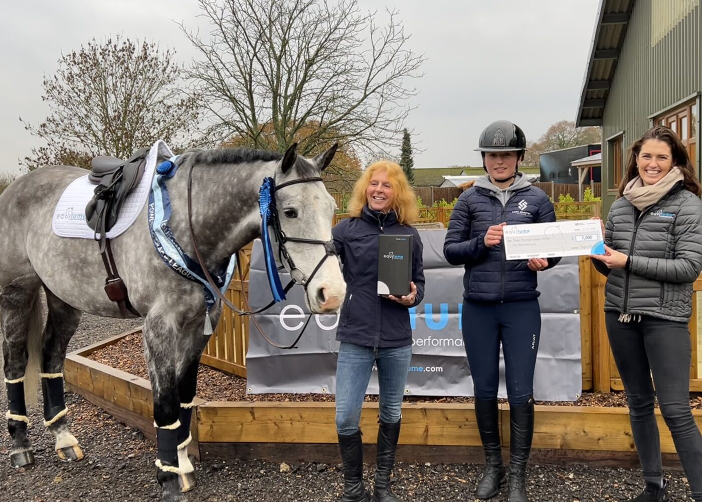 Grey horse wearing a saddle and bridle and his prize of a sash and a rosette on his head. Standing beside his owner Katie, rider Laura and Equilume marketing manager Alyssa O'Neill